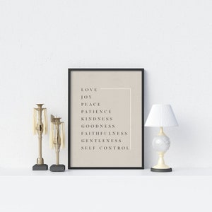 May include: A black framed print with a beige background and white text. The text reads "Love, Joy, Peace, Patience, Kindness, Goodness, Faithfulness, Gentleness, Self Control". The print is displayed on a white surface with two gold candle holders.