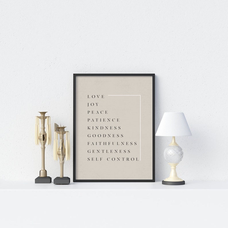 May include: A black framed print with a beige background and white text. The text reads "Love, Joy, Peace, Patience, Kindness, Goodness, Faithfulness, Gentleness, Self Control". The print is displayed on a white surface with two gold candle holders.