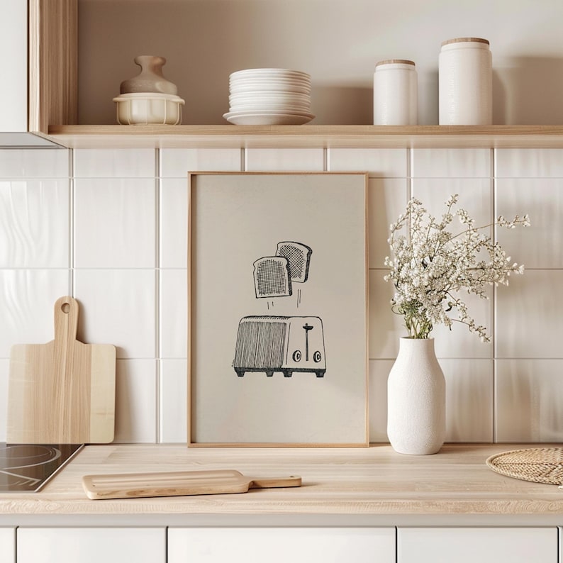 Puede incluir: Una ilustraci&oacute;n en blanco y negro de una tostadora con dos rebanadas de pan que salen disparadas. La ilustraci&oacute;n est&aacute; enmarcada y colgada en una pared de azulejos blancos en una cocina.