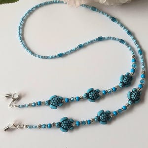 Op de afbeelding: Een lichtblauwe glazenketting met kleine, ronde en rechthoekige kralen. De ketting heeft vier turquoise schildpadbedels en zilverkleurige metalen sluitingen. De achtergrond omvat een witte bloem en een groen blad.