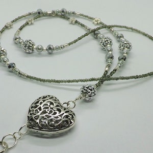 Puede incluir: Un charm de coraz&oacute;n plateado con un dise&ntilde;o intrincado cuelga de un cord&oacute;n de cuentas grises. El cord&oacute;n est&aacute; hecho de dos hebras de cuentas, una con cuentas plateadas y la otra con cuentas gris oscuro.