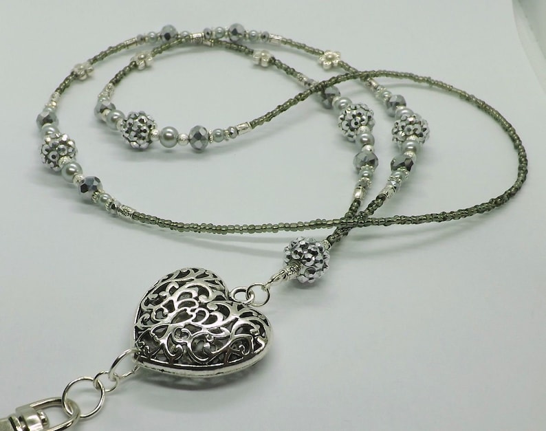 Puede incluir: Un charm de coraz&oacute;n plateado con un dise&ntilde;o intrincado cuelga de un cord&oacute;n de cuentas grises. El cord&oacute;n est&aacute; hecho de dos hebras de cuentas, una con cuentas plateadas y la otra con cuentas gris oscuro.