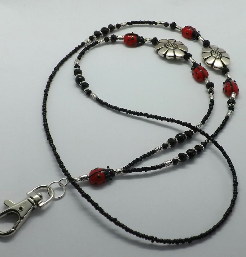 Pode incluir: Um cord&atilde;o de contas preto com detalhes de joaninhas vermelhas e flores prateadas. O cord&atilde;o tem um fecho prateado e foi projetado para um crach&aacute; de identifica&ccedil;&atilde;o ou chaves. As joaninhas s&atilde;o vermelhas com pintas pretas.
