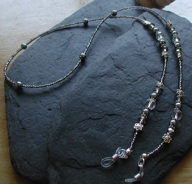 May include: A pair of black and silver beaded eyeglass chains with clear crystal beads and silver flower accents. The chains are attached to clear plastic eyeglass holders.