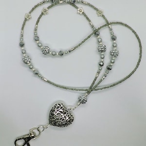 Puede incluir: Un charm de coraz&oacute;n plateado con un dise&ntilde;o intrincado cuelga de un cord&oacute;n de cuentas gris con detalles plateados. El cord&oacute;n tiene un cierre plateado.