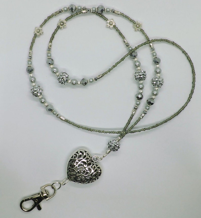 Puede incluir: Un charm de coraz&oacute;n plateado con un dise&ntilde;o intrincado cuelga de un cord&oacute;n de cuentas gris con detalles plateados. El cord&oacute;n tiene un cierre plateado.