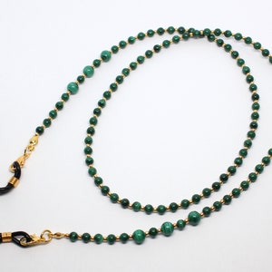 May include: A green and gold beaded eyeglass chain with black metal clasps. The chain is made of small, round beads with a gold finish. The beads are interspersed with larger, green malachite beads.