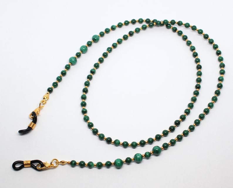 May include: A green and gold beaded eyeglass chain with black metal clasps. The chain is made of small, round beads with a gold finish. The beads are interspersed with larger, green malachite beads.