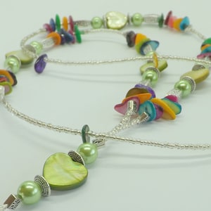 Op de afbeelding: Een meerstrengs ketting met groene, blauwe, oranje en paarse schelp kralen en zilveren accenten. De ketting heeft een hartvormige hanger.