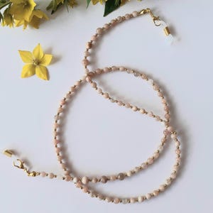 Puede incluir: Una cadena de gafas con pequeñas cuentas redondas de color melocotón y detalles dorados. La cadena está colocada sobre una superficie blanca, con flores amarillas en el fondo.