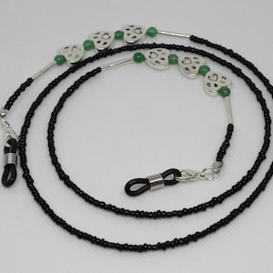 Puede incluir: Cadena de gafas de cuentas negras con dijes de corazón plateados y cuentas verdes. La cadena tiene dos extremos de goma negra con cierres metálicos.
