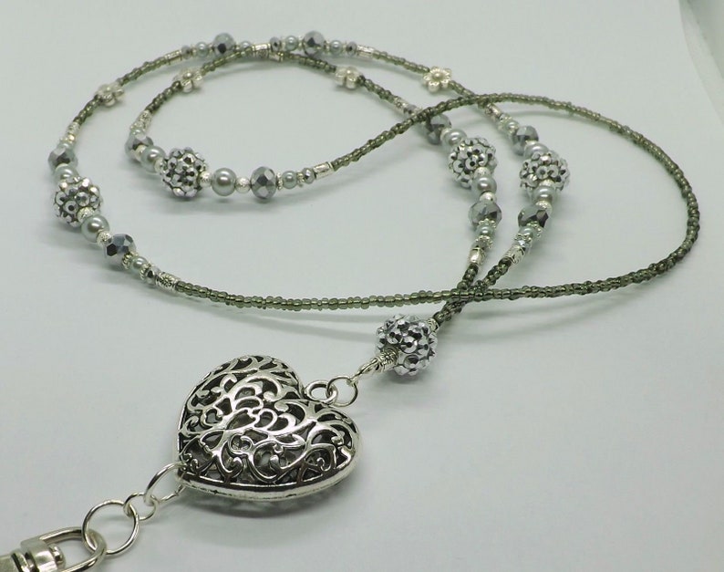 Puede incluir: Un charm de coraz&oacute;n plateado con un dise&ntilde;o intrincado cuelga de un cord&oacute;n de cuentas grises. El cord&oacute;n est&aacute; hecho de dos hebras de cuentas, una con cuentas plateadas y la otra con cuentas gris oscuro.