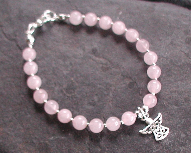 Może przedstawiać: Bransoletka z koralik&oacute;w z r&oacute;żowego kwarcu z zawieszką w kształcie anioła ze srebra. Bransoletka wykonana jest z okrągłych koralik&oacute;w i ma srebrną zapinkę.