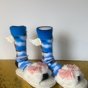 May include: A pair of white fuzzy slippers with pink bows. The slippers are worn with blue and white socks that have angel wings and the words "Little Sister" printed on them.