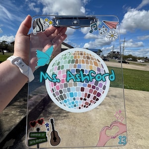 May include: Clear clipboard decorated with colorful stickers, including a disco ball, musical notes, a butterfly, and a guitar. The name "Ms. Ashford" is written in teal script. The clipboard is held up against a blue sky.