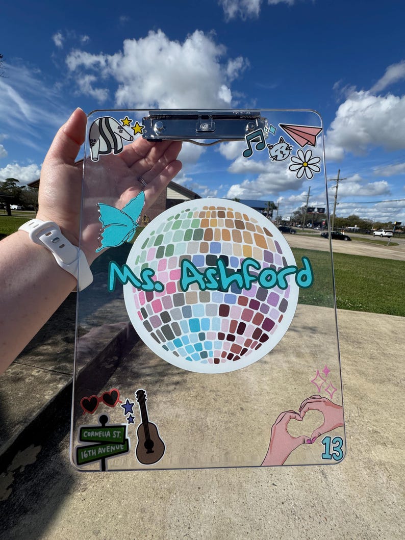 May include: Clear clipboard decorated with colorful stickers, including a disco ball, musical notes, a butterfly, and a guitar. The name "Ms. Ashford" is written in teal script. The clipboard is held up against a blue sky.