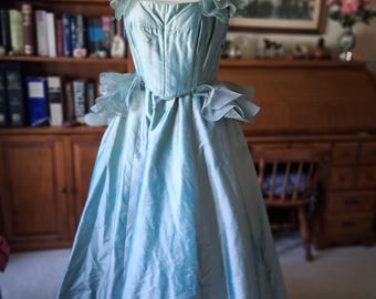 May include: A light blue vintage dress on a mannequin. The dress features a square neckline, ruffled cap sleeves, and a fitted bodice. The skirt is full and flowing, with a ruffled detail at the waist. The dress is made of a shiny, silky fabric.