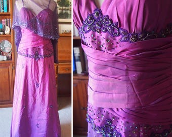 May include: A formal, floor-length purple gown with a sweetheart neckline and a matching sheer shawl. The dress features intricate beading and embellishments around the bodice and waist, with a flowing skirt. The dress is displayed on a mannequin.
