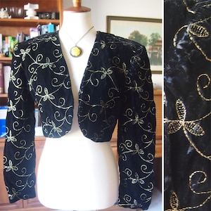 Può includere: Bolero in velluto nero con maniche lunghe e scollo a V. La giacca è adornata da motivi floreali ricamati in oro. Il bolero è esposto su un manichino, mostrando il suo design e la sua vestibilità. È visibile anche una collana con un pendente giallo.