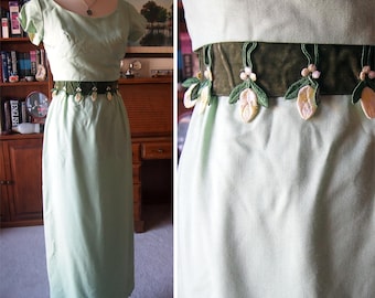May include: A light green vintage dress with short sleeves and a round neckline. The dress features a dark green velvet belt adorned with embroidered floral accents. The dress is displayed on a mannequin.