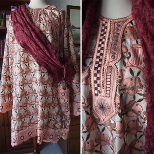May include: A long-sleeved, light-colored tunic with a floral pattern in shades of pink and brown. The tunic features intricate embroidery around the neckline and cuffs. A maroon scarf is draped over the shoulders. The garment is displayed on a mannequin.