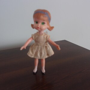 May include: A vintage plastic doll with brown hair, green eyes, and a light brown dress. The doll is wearing a blue headband and has a sweet expression.