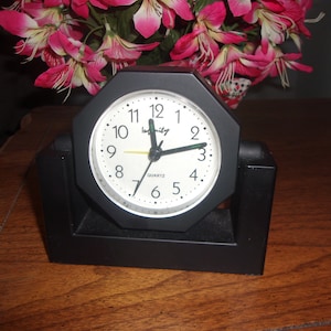May include: Black octagonal alarm clock with a white face and black hands. The clock has a green "Infinity" logo and says "Quartz" below the 6. The clock is sitting on a black stand.
