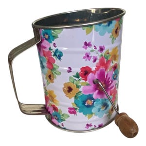 May include: A metal flour sifter featuring a white background with a vibrant floral pattern in shades of pink, teal, yellow, and green. The kitchen tool includes a silver metal handle, a silver rim, and a crank mechanism with a wooden knob.