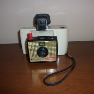 May include: A white Polaroid Swinger camera with a black lens and a red button. The camera is on a wooden surface with a black strap.