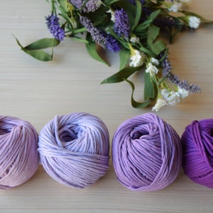 Puede incluir: Cuatro ovillos de hilo en tonos de morado, desde lavanda claro hasta violeta oscuro, dispuestos sobre una superficie de madera clara.