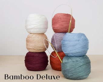 May include: A stack of six yarn balls in various colors, including white, peach, burgundy, orange, lavender, blue, and teal. A gold knitting needle is inserted into some of the yarn balls. The text "Bamboo Deluxe" is at the bottom.