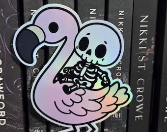 May include: A holographic sticker featuring a pink flamingo with a skeleton riding on its back. The flamingo has black accents and the skeleton is white. The sticker is placed in front of a row of books.