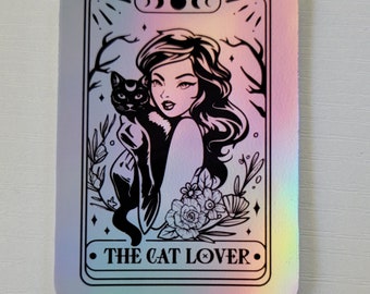 May include: A holographic sticker featuring a black and white illustration of a woman with long hair holding a black cat. The text "THE CAT LOVER" is printed below the image.
