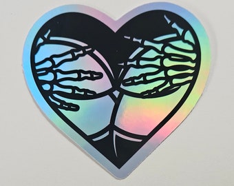 May include: A holographic sticker in the shape of a heart with a black outline. The heart features two skeletal hands clasped together.