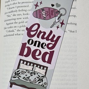 May include: A pink and white bookmark with a bed and the text "Only one bed" in a dark red font. The bookmark also features a small pink and white motel sign with the text "The Bookish Inn" and a key with a heart-shaped charm.