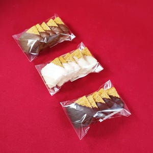 May include: Three clear plastic bags containing triangular wheat cereal snacks. One bag features milk chocolate coating, one features white chocolate coating, and one features dark chocolate coating, each with visible drizzled chocolate lines.