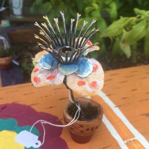May include: A small, hand-painted flower sculpture with a metal center and red, blue, and yellow petals. The flower is in a small terracotta pot with a brown tag.