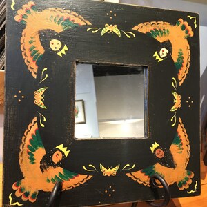 May include: A black square mirror with a painted frame. The frame features a stylized design of birds with yellow, orange, and green accents. The mirror is on a stand with two black metal hooks.