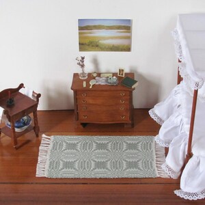 Handmade, Handcrafted 1:12 Scale Dollhouse Miniature Rug, Artisan Hand Woven Jade Green Carpet for Traditional Country Farmhouse or Modern Coastal Beach Cottage Decor is a perfect Doll House Furniture Accessory for your Miniature Bedroom or Living Room! Shown in a Bedroom scene with white canopy bed with a white ruffled bed skirt and bedspread, wooden dresser with  a small mirror, a book, and a few decorative items, and a wash table. A framed landscape picture is on the wall.