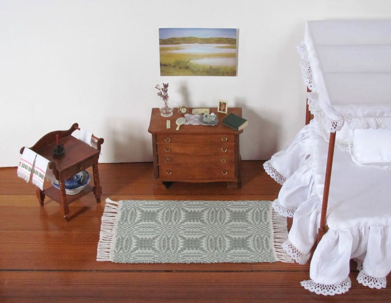 Handmade, Handcrafted 1:12 Scale Dollhouse Miniature Rug, Artisan Hand Woven Jade Green Carpet for Traditional Country Farmhouse or Modern Coastal Beach Cottage Decor is a perfect Doll House Furniture Accessory for your Miniature Bedroom or Living Room! Shown in a Bedroom scene with white canopy bed with a white ruffled bed skirt and bedspread, wooden dresser with  a small mirror, a book, and a few decorative items, and a wash table. A framed landscape picture is on the wall.