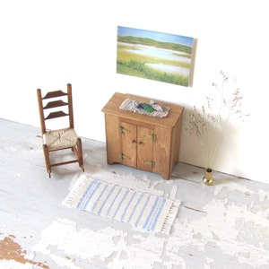 May include: A miniature wooden cabinet with two doors, a small rug, a rocking chair, and a framed landscape painting on a white wall.