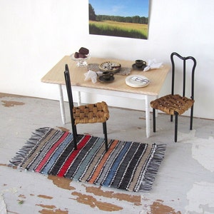May include: A miniature dining set with a wooden table, two chairs with woven seats, and a striped rug. The table is set with a plate, a cup, and a knife.