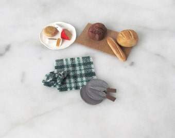 May include: A miniature picnic scene featuring a white plate with cheese and bread, a green checked cloth, two small grey chopping boards, and a brown wooden board with two loaves of bread.