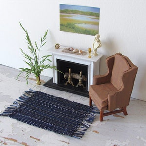 Handcrafted, Inch to Foot Dollhouse Miniature Rug is 1:12 Scale Artisan Hand Loom Woven in Indigo Navy Ocean Blue, Beige, and White Stripe Floor Carpet for Modern Coastal Seaside Nautical Beach House, Cabin Decor Doll House Furniture Accessory. Styled with a chair in front of a fireplace, with a green plant in a floor vase and marsh landscape.