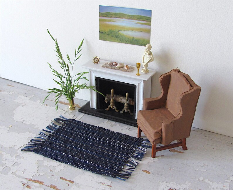Handcrafted, Inch to Foot Dollhouse Miniature Rug is 1:12 Scale Artisan Hand Loom Woven in Indigo Navy Ocean Blue, Beige, and White Stripe Floor Carpet for Modern Coastal Seaside Nautical Beach House, Cabin Decor Doll House Furniture Accessory. Styled with a chair in front of a fireplace, with a green plant in a floor vase and marsh landscape.