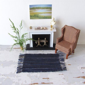 Handcrafted, Inch to Foot Dollhouse Miniature Rug is 1:12 Scale Artisan Hand Loom Woven in Indigo Navy Ocean Blue, Beige, and White Stripe Floor Carpet for Modern Coastal Seaside Nautical Beach House, Cabin Decor Doll House Furniture Accessory. Styled with a chair and couch in front of a fireplace, with a green plant in a floor vase and marsh landscape.