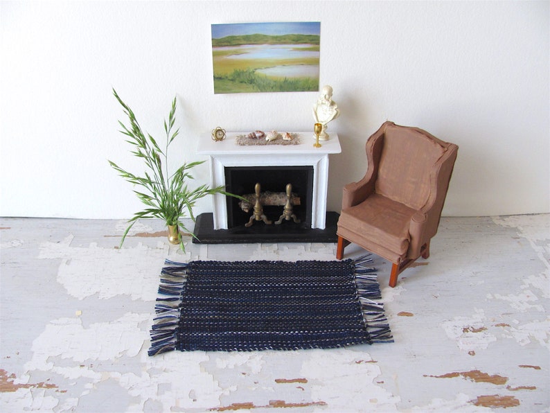 Handcrafted, Inch to Foot Dollhouse Miniature Rug is 1:12 Scale Artisan Hand Loom Woven in Indigo Navy Ocean Blue, Beige, and White Stripe Floor Carpet for Modern Coastal Seaside Nautical Beach House, Cabin Decor Doll House Furniture Accessory. Styled with a chair and couch in front of a fireplace, with a green plant in a floor vase and marsh landscape.