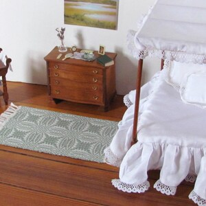 Handmade, Handcrafted 1:12 Scale Dollhouse Miniature Rug, Artisan Hand Woven Jade Green Carpet for Traditional Country Farmhouse or Modern Coastal Beach Cottage Decor is a perfect Doll House Furniture Accessory for your Miniature Bedroom or Living Room! Shown in a Bedroom scene with white canopy bed with a white ruffled bed skirt and bedspread, wooden dresser with  a small mirror, a book, and a few decorative items, and a wash table. A framed landscape picture is on the wall.