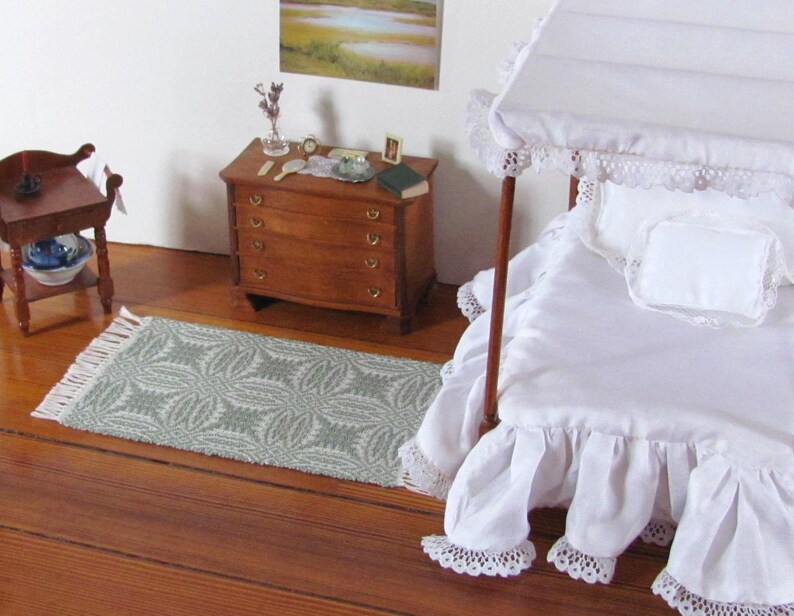 Handmade, Handcrafted 1:12 Scale Dollhouse Miniature Rug, Artisan Hand Woven Jade Green Carpet for Traditional Country Farmhouse or Modern Coastal Beach Cottage Decor is a perfect Doll House Furniture Accessory for your Miniature Bedroom or Living Room! Shown in a Bedroom scene with white canopy bed with a white ruffled bed skirt and bedspread, wooden dresser with  a small mirror, a book, and a few decorative items, and a wash table. A framed landscape picture is on the wall.