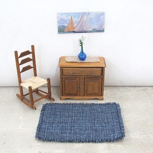 Inch to Foot Dollhouse Miniature Rug, 1:12 Scale Woven Small Indigo Navy Blue and Light Blue Floor Carpet for Modern Coastal Seaside Cottage or Traditional Rustic Nautical Beach Doll House. Styled with a rocking chair, cabinet with a linen runner and blue glass vase with white flowers, a picture of racing sailing ships on the wall.